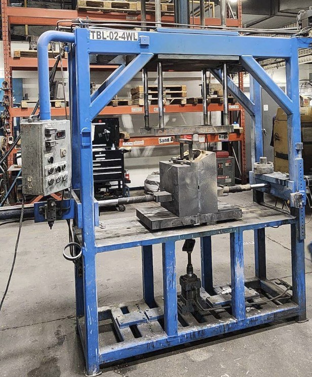 The four-way casting table, a blue steel frame with a steel permanent mold sitting on the deck, hydraulic actuator rods entering from either side, and a pushbutton enclosure bolted to the frame