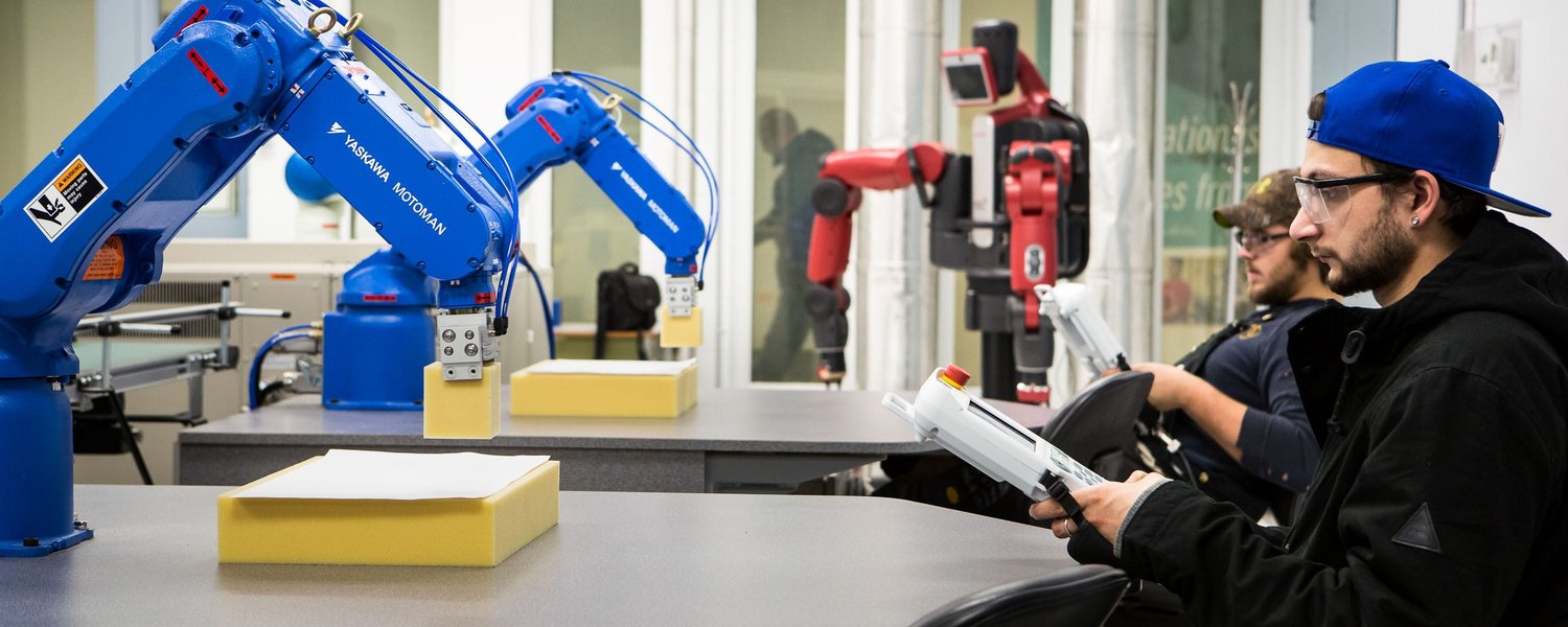 Robotics lab with two blue Yaskawa Motoman six-axis arms on benches and a student holding a teach pendant
